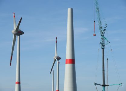 Workers standing in a basket hang from a crane next to a wind turbine under construction.