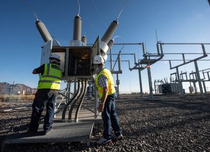 Two utility workers in yellow vests work on a transmission system